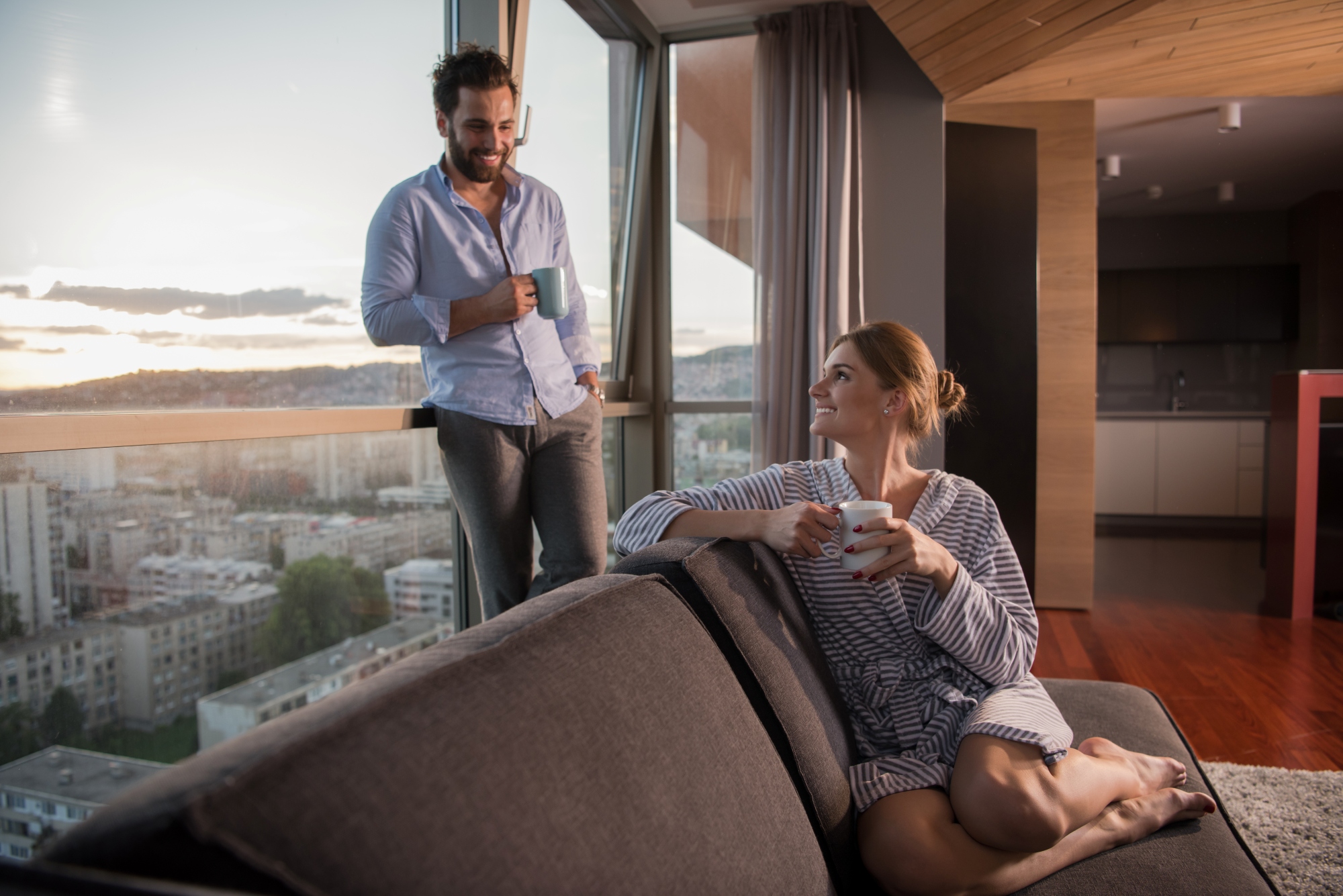 pareja-joven-romantica-y-feliz-disfrutando-del-cafe-de-la-tarde-y-el-hermoso-paisaje-de-la-puesta-de-sol-de-la-ciudad-mientras-esta-de-pie-junto-la-ventana