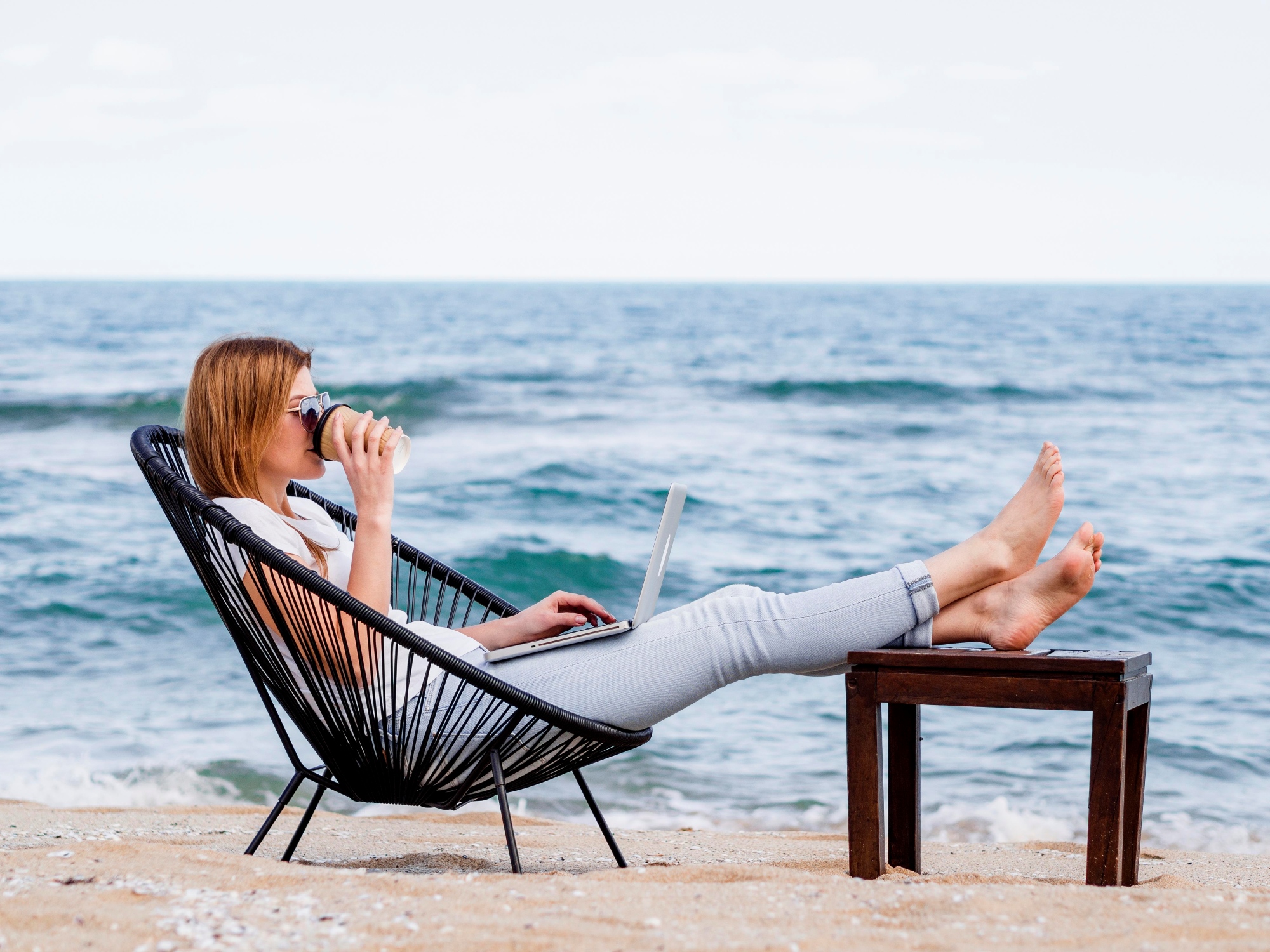 Mujer tomando café en la playa mientras disfruta de la brisa marina su trabajo en computadora portátil frente al mar