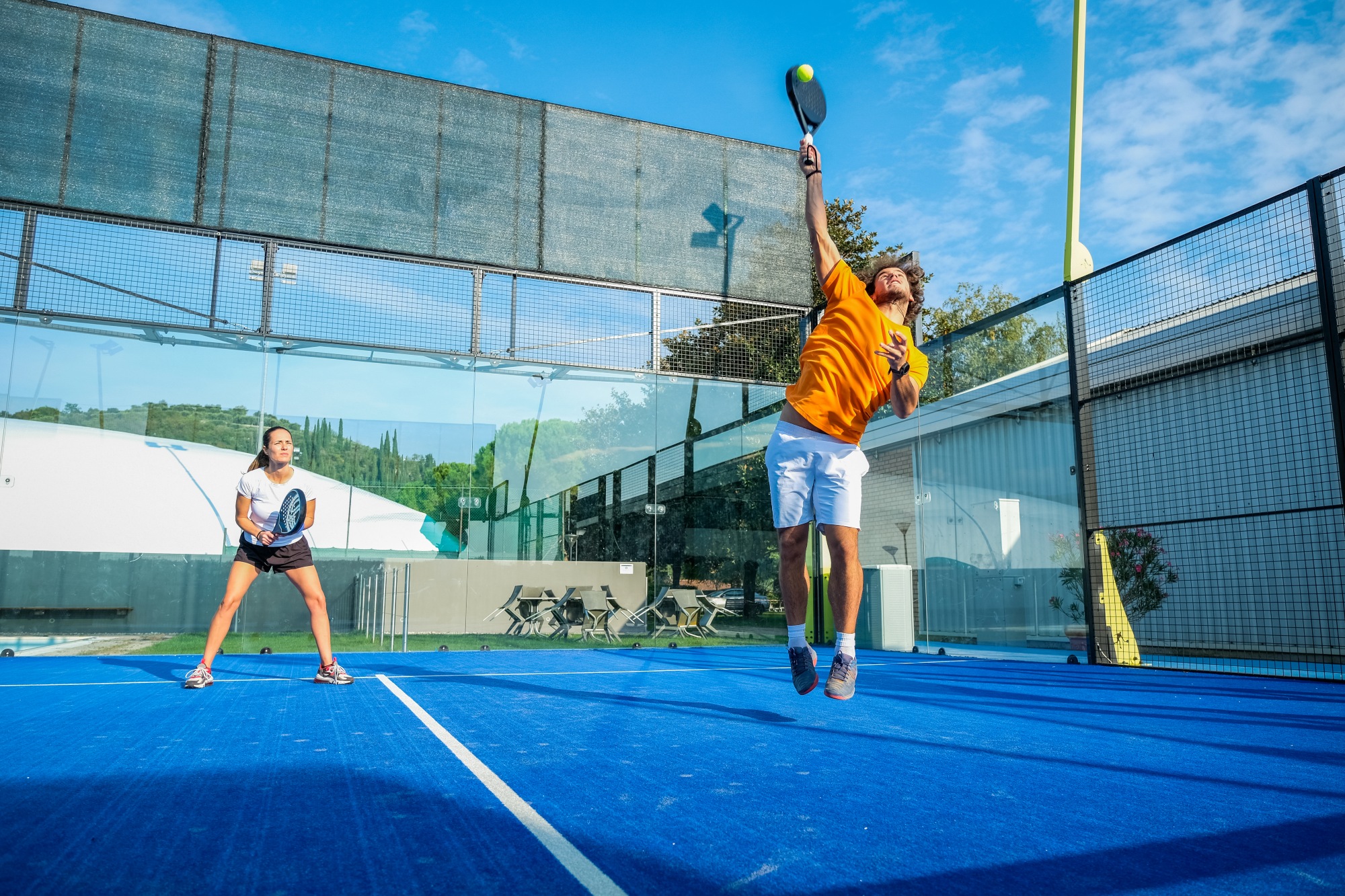 Pareja hombre mujer jugando padel hombre da un gran salto para poder devi¿olver la pelotas a los contricantes mujer al fondo de la cncha cubriendo su area de juego